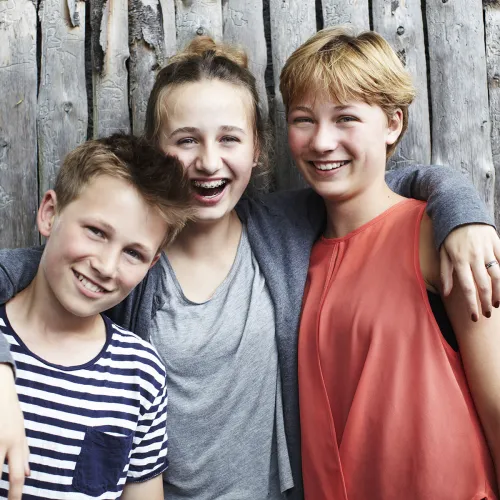 A girl puts her arms around her sister and brother as all three stand laughing as they pose for a picture